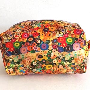 May include: A rectangular fabric pouch featuring a vibrant, multicoloured pattern of concentric circles and dots in shades of red, orange, yellow, green, blue, and purple. The bag has a zip closure and a small white tag labelled HAND MADE.