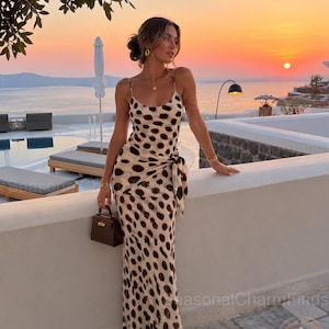 Könnte beinhalten: Ein bodenlanges ärmelloses Kleid mit cremefarbener Basis und großen dunkelbraunen Punkten, einem Rundhalsausschnitt, dünnen Spaghettiträgern und einer seitlichen Bindung an der Taille. Das Kleid wird mit einer kleinen dunkelbraunen Handtasche kombiniert.