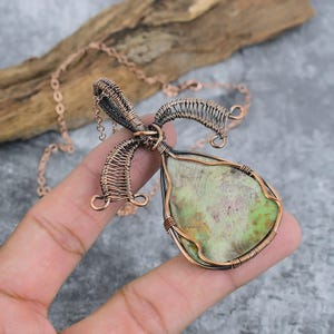 May include: A teardrop-shaped pendant featuring a mottled light green and brown stone encased in intricate copper wire wrapping. The pendant is attached to a matching copper chain, with decorative wire loops extending from the top bail.
