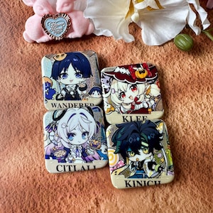 May include: Four square pinback buttons featuring chibi-style anime characters with holographic backgrounds. The buttons are labeled with the names WANDERER, KLEE, CITLALI, and KINICH in black text on a cream-colored banner at the bottom of each.