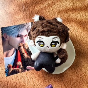May include: A plush doll with brown hair, white ear tufts, and yellow eyes, wearing a black sleeveless top. Beside it is a rectangular card featuring an illustration of a person with silver hair and a cat, set against a textured orange background.
