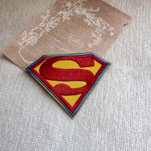 May include: A diamond-shaped embroidered patch featuring the iconic Superman logo with a bold red letter S centered on a bright yellow background, enclosed by a thick red border and a thin blue outer edge, resting on a tan card with floral art.