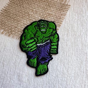 Puede incluir: Un parche bordado que muestra a un personaje musculoso de color verde con una expresión facial severa, vistiendo pantalones morados. El personaje aparece en una postura inclinada hacia adelante con los puños cerrados y delineado en hilo negro.