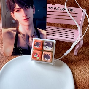 May include: A square white mechanical keyboard keycap display base featuring four individual keycaps with cartoon character illustrations, including one with an apple motif. The display is accompanied by a small pink wooden bench ornament.
