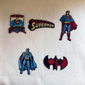 以下が含まれることがあります： コミックのキャラクターやロゴが刺繍された5つの布製ワッペン。テキスト入りのスーパーマンの盾ロゴ、スーパーマンの文字ワッペン、スーパーマンの全身フィギュア、バットマンの全身フィギュア、黒と赤のコウモリ型のエンブレムが含まれています。