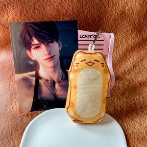 May include: A rectangular plush keychain featuring a yellow and orange cat-like character with a smiling face, attached to a metal ring. Behind it stands a portrait card of a young person with dark hair and a necklace, resting on a pink stand.