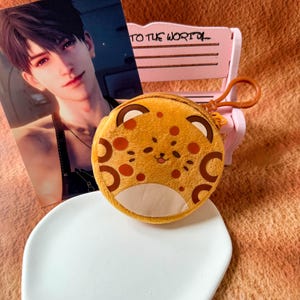 May include: A round, plush, yellow leopard-patterned coin purse with a small brown plastic keychain attachment. The purse features a cartoon face with whiskers, a small tongue, and dark brown spots. It is positioned in front of a pink bench.