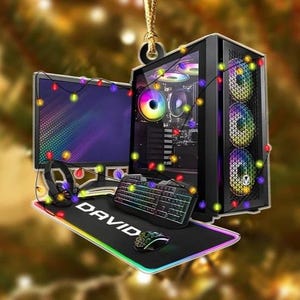May include: A hanging ornament featuring a gaming computer setup with a monitor, a tower case with RGB fans, a keyboard, a mouse, and a mousepad with the name DAVID printed on it. The entire setup is wrapped in a string of colorful Christmas lights.