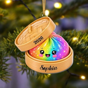 Könnte beinhalten: Ein hängendes Ornament in Form eines Bambusdampfkorbs, das einen regenbogenfarbenen, glitzernden Knödel mit lächelndem Gesicht enthält. Der Deckel zeigt das Jahr 2026 und der Boden ist mit dem Namen Sophia in schwarzer Schreibschrift personalisiert.