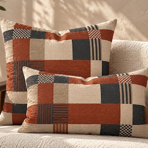 Könnte beinhalten: Zwei quadratische und rechteckige Dekokissen mit einem Patchwork-Design aus Stoffblöcken in Rostorange, Anthrazit und Creme. Das Muster besteht aus einfarbigen Flächen, feinen vertikalen Streifen und kleinen karierten Quadraten in Webstruktur.