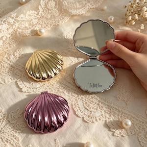 May include: Three shell-shaped compact mirrors in gold, pink, and silver. One silver mirror is open, revealing a reflective surface with the name Tabitha engraved in script. The mirrors feature a scalloped edge design and a metallic finish.
