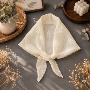 May include: A triangular off-white headscarf featuring delicate white floral embroidery scattered across the fabric. The edges of the scarf are finished with a decorative scalloped lace trim, and the fabric is tied in a knot at the centre front.