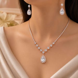 Könnte beinhalten: Ein Set aus einer roségoldenen Halskette und passenden Ohrhängern mit klaren, facettierten Kristallen. Die Kette besteht aus einer feinen Reihe runder Kristalle, die in einen tropfenförmigen Anhänger münden, ergänzt durch tropfenförmige Ohrringe.