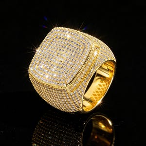 May include: A gold-toned ring featuring a large, square-shaped face completely encrusted with numerous small, clear, sparkling stones. The band and the sides of the square setting are also densely paved with the same clear, reflective stones.