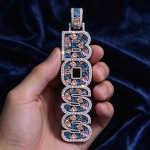 May include: A vertical pendant featuring the word BOSS in block letters, encrusted with small clear stones and inlaid with a mosaic of light pink and deep blue pear-shaped and rectangular stones, finished with a matching stone-set bail.