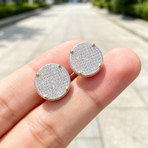 May include: A pair of circular stud earrings featuring a gold-toned metal frame with a grid-like setting. Each earring is densely encrusted with numerous small, clear, sparkling stones that cover the entire flat surface of the round disc design.