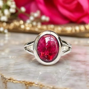 Puede incluir: Un anillo de plata con una gran piedra preciosa de color rosa intenso en forma ovalada, engastada en un bisel liso. La banda del anillo tiene un diseño dividido, ramificándose en dos segmentos de plata separados a cada lado de la piedra.