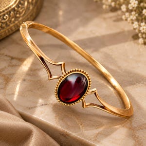 Peut inclure: Un bracelet jonc articulé doré orné d'une pierre précieuse ovale rouge foncé en son centre. La pierre est sertie dans un cadre doré perlé, flanquée de détails métalliques géométriques ajourés reliant le bracelet lisse et poli.
