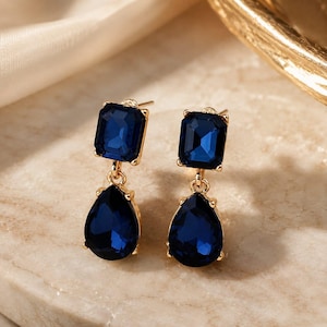 May include: A pair of gold-tone drop earrings featuring a rectangular dark blue faceted stone at the top and a larger pear-shaped dark blue faceted stone suspended below. The stones are set in gold-colored metal frames with secure post backings.