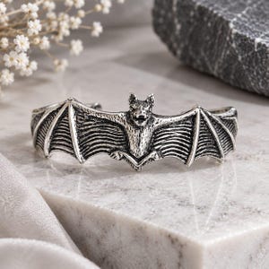 May include: A silver-toned metal cuff bracelet featuring a detailed bat with outstretched wings. The bat is centered on the band, displaying textured wing membranes and a sculpted head with an open mouth. The metal has an oxidized finish.
