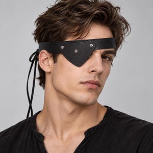 Op de afbeelding: Een zwarte leren ooglap in een driehoekige vorm met drie zilverkleurige metalen klinknagels. De ooglap is bevestigd aan een dunne zwarte leren hoofdband die om het hoofd loopt en aan de zijkant wordt vastgemaakt met dunne leren koorden.