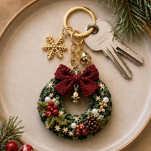 May include: A gold-toned metal keychain featuring a crocheted dark green wreath adorned with a deep red fabric bow, a small gold bell, white pearl beads, red berries, and a brown pinecone. A gold snowflake charm hangs from the ring.