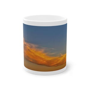 Może przedstawiać: Biały ceramiczny kubek z fotograficznym nadrukiem nieba o zachodzie słońca. Obraz przedstawia gradient od głębokiego błękitu u góry, przechodzący w żywe pomarańczowe i złotożółte chmury rozciągające się poziomo przez środek kubka.