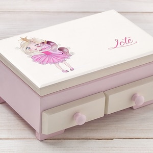 Puede incluir: Una caja de joyería de madera rosa con dos cajones y una tapa blanca. La tapa presenta una ilustración de dibujos animados de una niña con cabello rubio que lleva una corona y un vestido rosa abrazando a un unicornio. El nombre "Jote" está escrito en cursiva rosa en la tapa.