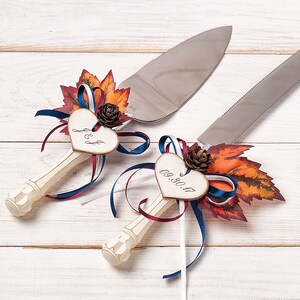 May include: A pair of white cake servers with decorative heart-shaped tags. The tags are decorated with fall leaves, ribbons, and pine cones. One tag says "J & J" and the other says "09.30.17".