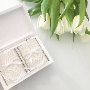 Pu&ograve; includere: Scatola portafedi bianca con due cuscini portafedi in raso bianco con bordo in pizzo. La scatola &egrave; aperta e i cuscini sono visibili. La scatola &egrave; appoggiata su una superficie bianca. Sullo sfondo ci sono dei tulipani bianchi.