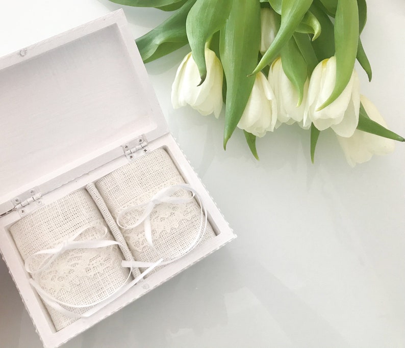 Pu&ograve; includere: Scatola portafedi bianca con due cuscini portafedi in raso bianco con bordo in pizzo. La scatola &egrave; aperta e i cuscini sono visibili. La scatola &egrave; appoggiata su una superficie bianca. Sullo sfondo ci sono dei tulipani bianchi.