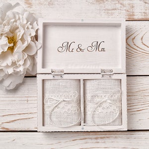 Pu&ograve; includere: Scatola portafedi in legno bianco con due scomparti rivestiti in tessuto bianco e pizzo. Il coperchio della scatola ha incisa la scritta "Mr. & Mrs.".