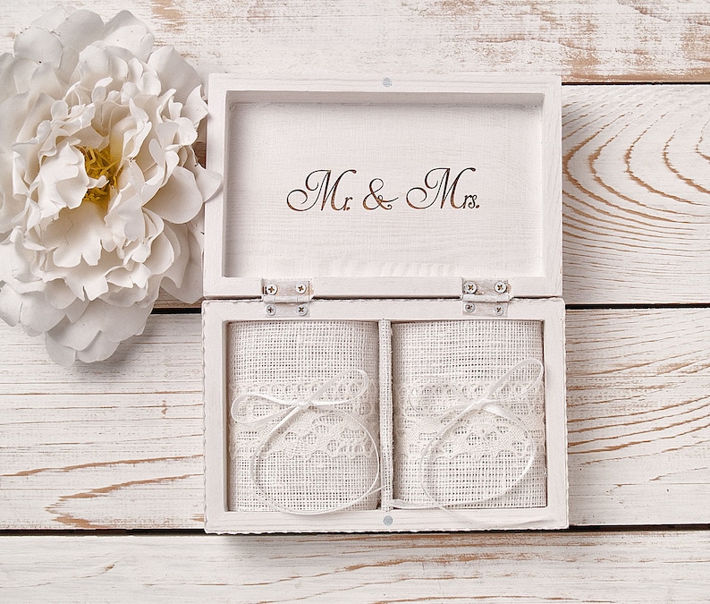 Pu&ograve; includere: Scatola portafedi in legno bianco con due scomparti rivestiti in tessuto bianco e pizzo. Il coperchio della scatola ha incisa la scritta "Mr. & Mrs.".