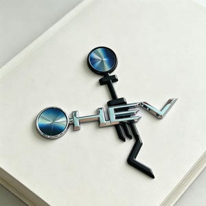 Peut inclure: Un emblème métallique arborant les lettres HEV en finition chromée, intégré à deux accents circulaires présentant un dégradé radial bleu et argent. Le design comprend une structure noire avec une forme géométrique verticale et angulaire.