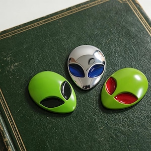 Op de afbeelding: Drie alien-hoofdspelden. Eén speld is zilverkleurig met blauwe ogen, één is limoengroen met zwarte ogen en één is limoengroen met rode ogen. Elke speld heeft een glad, afgerond, ovaalvormig ontwerp met opvallende, amandelvormige ooguitsparingen.