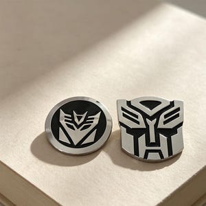 May include: Two metal pins with a polished silver and black finish. One pin is circular and features the Decepticon logo, while the other pin is shaped like the Autobot logo. Both emblems are rendered in a clean, high-contrast metallic design.