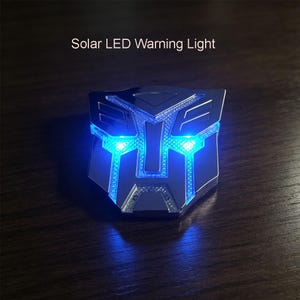 May include: A silver-coloured Solar LED Warning Light shaped like a robotic face emblem. The device features a metallic finish with integrated blue LED lights that illuminate the textured sections of the design. The text Solar LED Warning Light is visible.