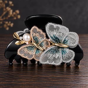 May include: A black hair claw clip featuring two embroidered butterfly appliqués. The left butterfly is accented with a white faux pearl and gold-toned metal leaves, while both butterflies display intricate teal, silver, and gold thread detailing.