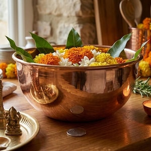 May include: A large, polished copper bowl filled with vibrant orange and yellow marigold flowers and green leaves. The bowl sits on a wooden surface alongside a small copper candle holder with a lit flame and a brass tray holding small figurines.