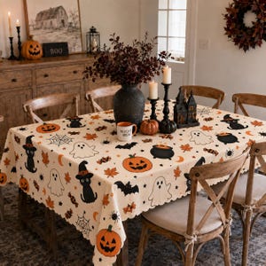 May include: A cream-colored tablecloth featuring a repeating Halloween pattern of orange jack-o'-lanterns, black cats wearing witch hats, white ghosts, black bats, spiders, spiderwebs, orange autumn leaves, and black cauldrons with orange contents.