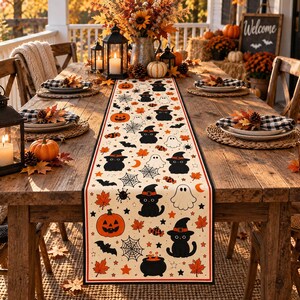 May include: A rectangular table runner featuring a repeating Halloween pattern on a cream background. The design includes black cats wearing orange hats, orange jack-o'-lanterns, white ghosts, black bats, spider webs, and orange autumn leaves.
