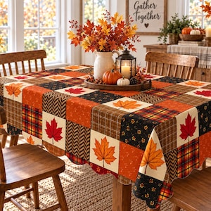Puede incluir: Un mantel rectangular con un diseño de patchwork de cuadrados con temática otoñal en tonos naranja, rojo, marrón y negro. El patrón incluye estampados de hojas de arce, cuadros, chevrón y motivos florales con un borde festoneado.