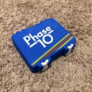 May include: A blue rectangular plastic storage case for the card game Phase 10. The lid features the white text Phase 10 with a stylized graphic design consisting of a thin yellow line and a thicker red line running diagonally across the bottom right corner.