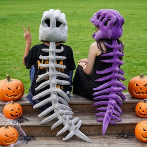 May include: Two plush skeleton headpieces with long, segmented spinal cord tails. One headpiece is light gray, and the other is vibrant purple. Both feature skull-like designs with hollow eye sockets and rib-like structures extending down the back.