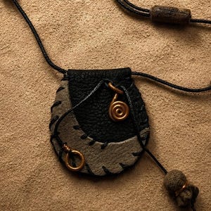 Puede incluir: Un collar con bolsa de cuero en negro y gris que incluye un cordón largo negro con cuatro cuentas cilíndricas marrones. La bolsa tiene un aplique de cuero gris en forma de media luna sobre una base negra texturizada con un pequeño dije de espiral dorado.