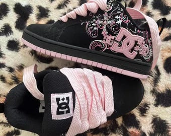 May include: A pair of black and light pink DC sneakers featuring thick light pink laces, a black suede-like upper with white and pink swirling graphic patterns, a pink DC logo on the side, and a silver-toned cross charm attached to the eyelets.