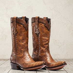 Può includere: Un paio di stivali da cowboy in pelle color cuoio con un motivo testurizzato a pelle di coccodrillo sul piede e sul tallone, con gambali in pelle liscia decorati da intricate cuciture e profili marrone scuro lungo le cuciture.