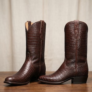 Op de afbeelding: Een paar donkerbruine cowboylaarzen met een textuur van krokodillenleer. De laarzen hebben een hoge schacht, een puntige neus, een hak en treklussen aan de bovenkant van de schacht. Het leer heeft een diepe, rijke bruine afwerking.