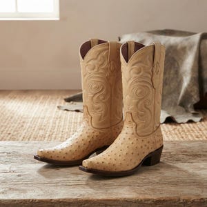 Op de afbeelding: Een paar lichtbeige cowboylaarzen met een textuurpatroon op de voet en gedetailleerde stiksels in dezelfde kleur op de hoge schacht. De laarzen hebben een puntige neus, een hak en een donkerbruine zool, wat zorgt voor een klassiek model.