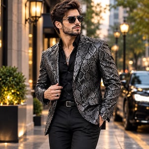 Puede incluir: Una americana con un patrón de piel de serpiente en plata metálica y negro, sobre una camisa negra abotonada y pantalones negros. La americana tiene solapas de muesca, dos bolsillos delanteros con solapa y cierre de un solo botón.